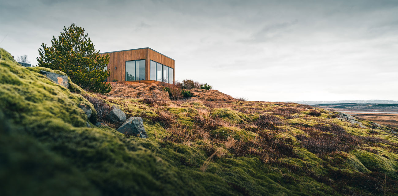 An image of a timber building on the edge of a green, rocky cliff with the beach and sea in the background of a grey day.